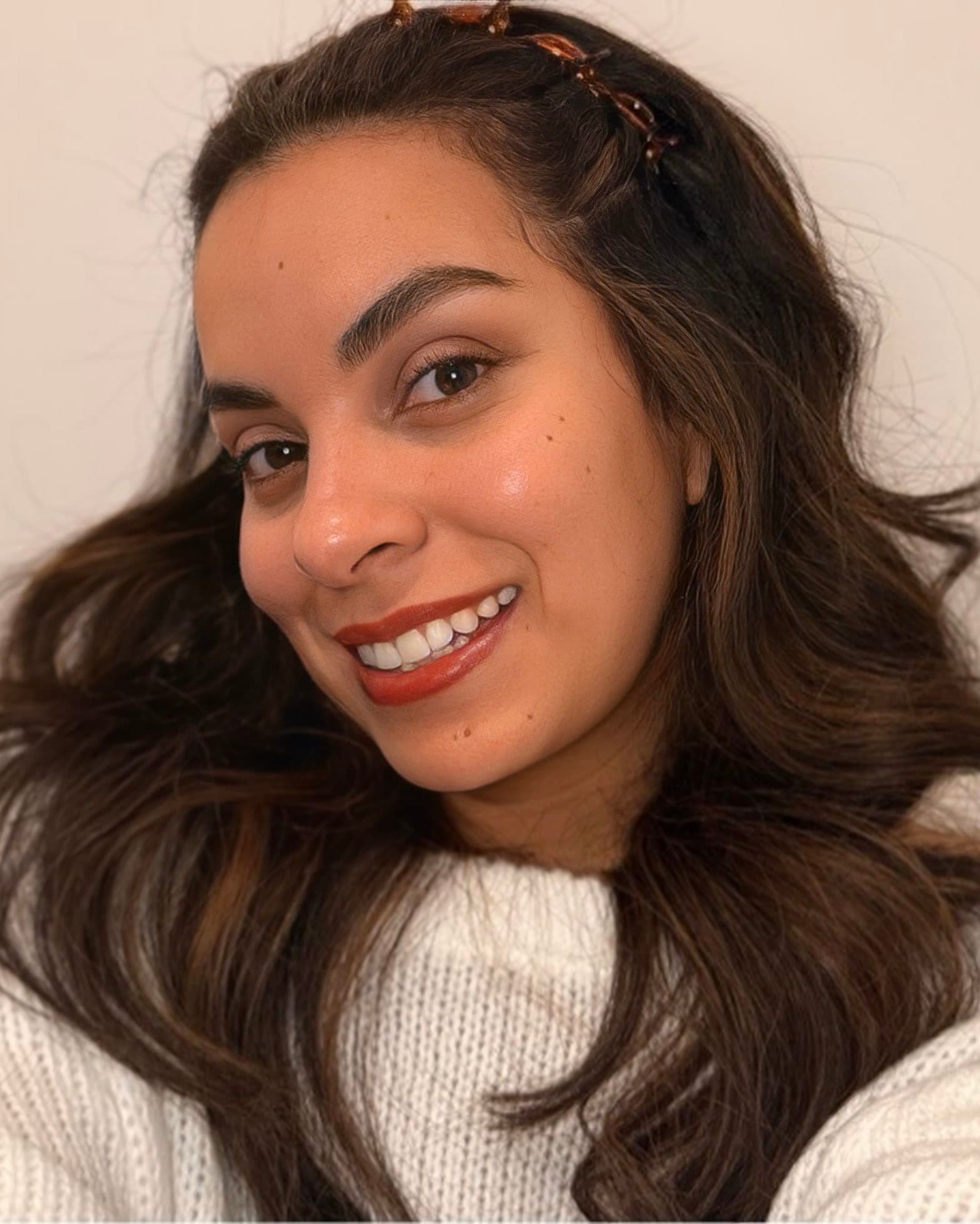 Retrato de Lorena: sonrisa abierta y cercana, mirando a cámara, con suéter de punto crema, broches en el pelo y labios terracota, bajo una luz cálida y suave.